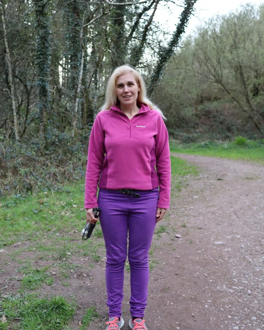 Sinead standing on a dirt trail in a wooded area surrounded by plants and trees. Sinead is dressed in purple